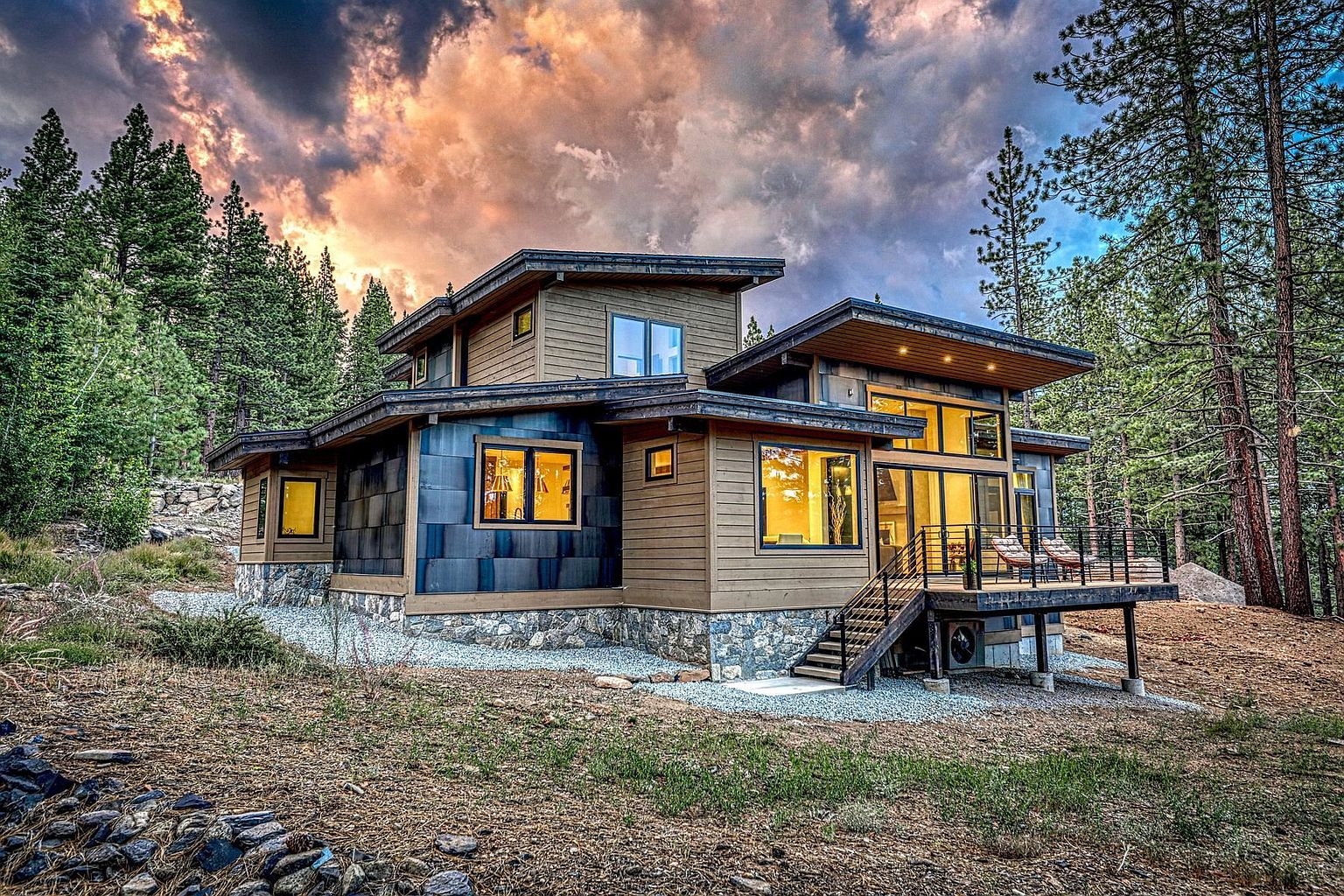 This contemporary mountain home features a striking mix of horizontal wood siding, dark metal paneling, and a stone foundation. The rear elevation showcases a raised deck with a metal railing, large windows that illuminate the interior, and a dramatic roofline set against a vibrant, cloudy sunset. The surrounding landscape consists of a natural, wooded forest setting with a gravel path leading toward the structure.