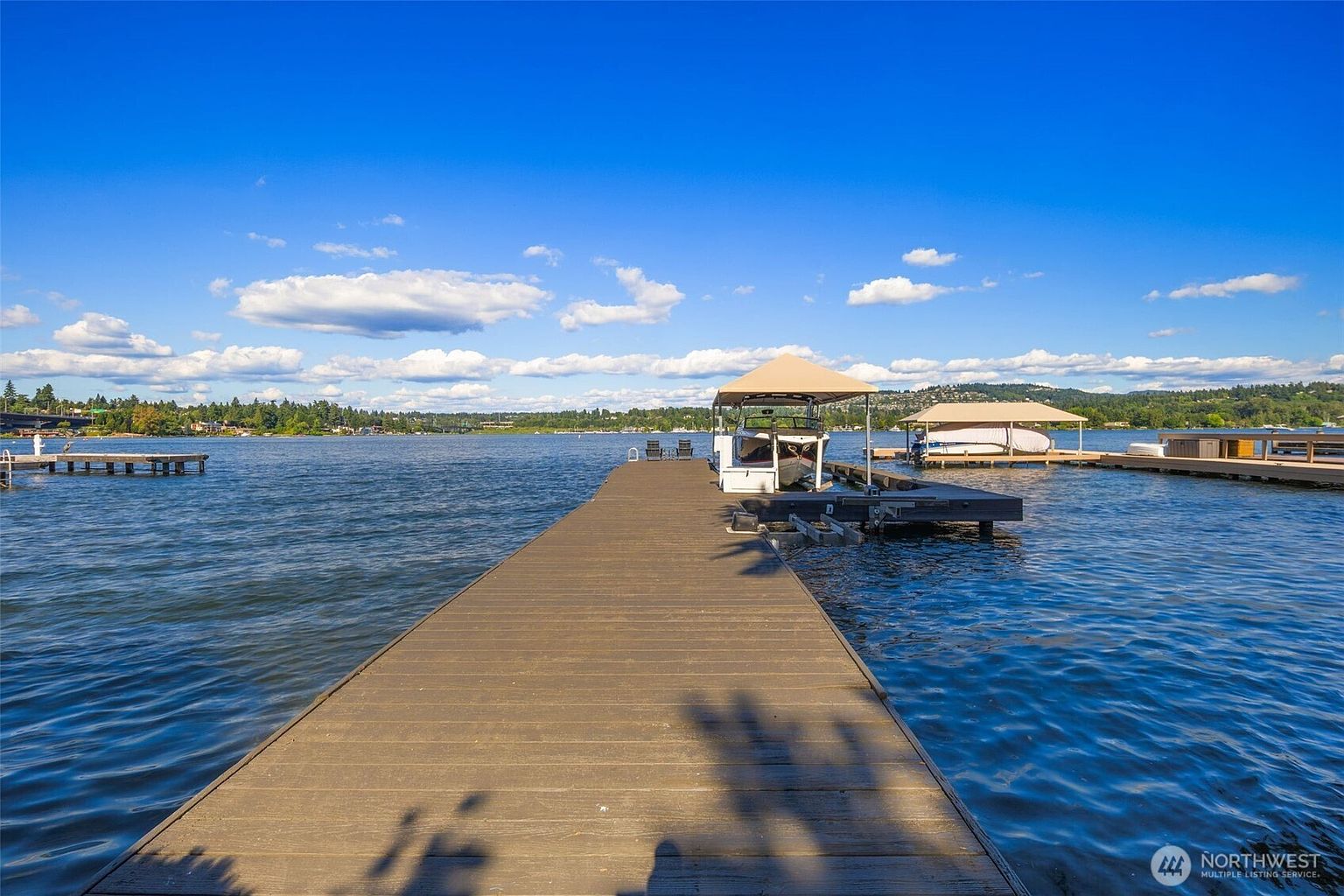 This perspective captures a long, wooden dock extending out into a calm, expansive lake under a bright blue sky with scattered clouds. The dock features a covered boat slip with a boat moored inside, alongside a second covered slip nearby, highlighting the property's direct water access. The scene offers a serene, recreational atmosphere, showcasing the waterfront lifestyle and the surrounding natural landscape.