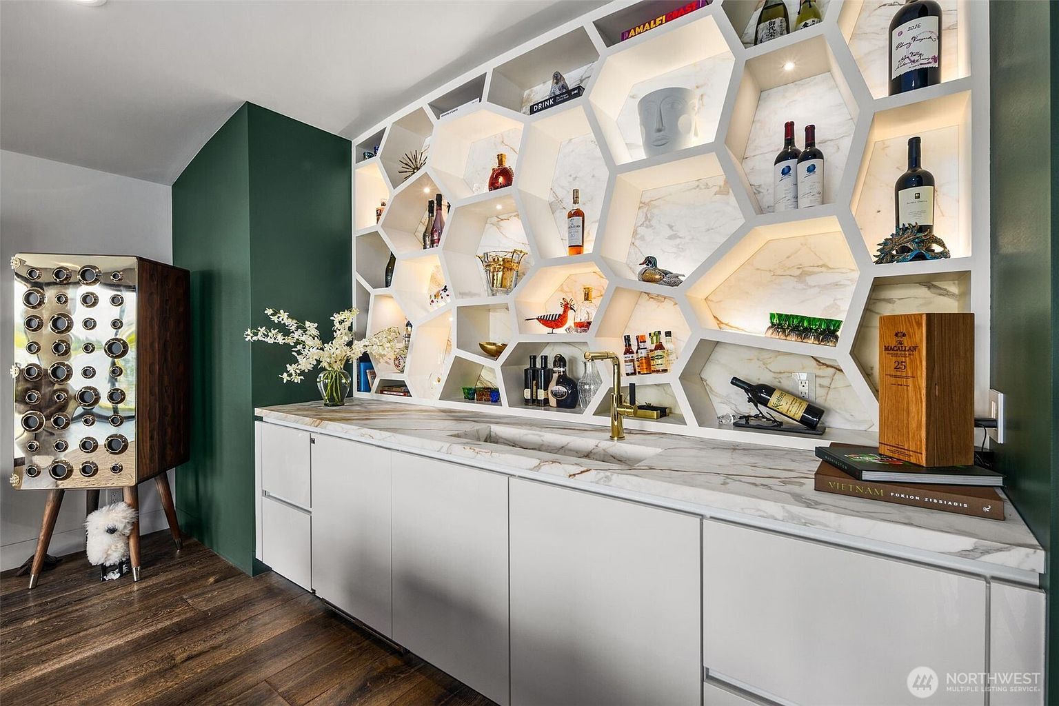 This sophisticated wet bar area features a unique honeycomb-patterned shelving unit with integrated lighting, set against a stone-patterned backsplash. The space includes a sleek, light-toned countertop with an undermount sink and gold-toned faucet, complemented by dark green walls and a modern, metallic-finished wine storage cabinet to the left. The composition is captured from a slightly elevated eye-level perspective, emphasizing the high-end finishes and curated display of spirits and glassware.