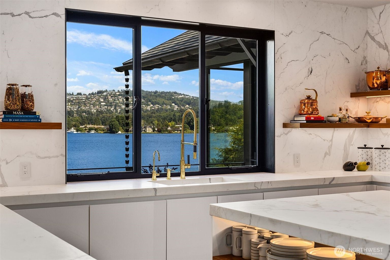This modern kitchen features a large window overlooking a serene lake and hillside, framed by clean white stone surfaces and minimalist cabinetry. A gold-toned faucet stands out against the backdrop, while open shelving displays curated decor and kitchenware. The bright, airy atmosphere is enhanced by the natural light pouring in, creating a sophisticated and tranquil culinary space.