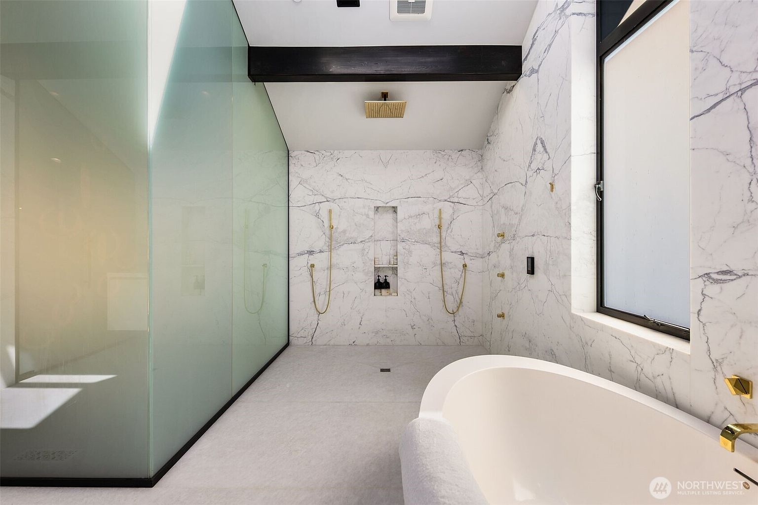 This luxurious primary bathroom features a spacious walk-in shower area with a large, frosted glass partition and a striking stone-clad wall with dual shower heads. A freestanding soaking tub sits in the foreground, complemented by gold-toned fixtures and a dark wood ceiling beam that adds architectural contrast. The space is brightened by a large window, creating a serene and high-end spa-like atmosphere.
