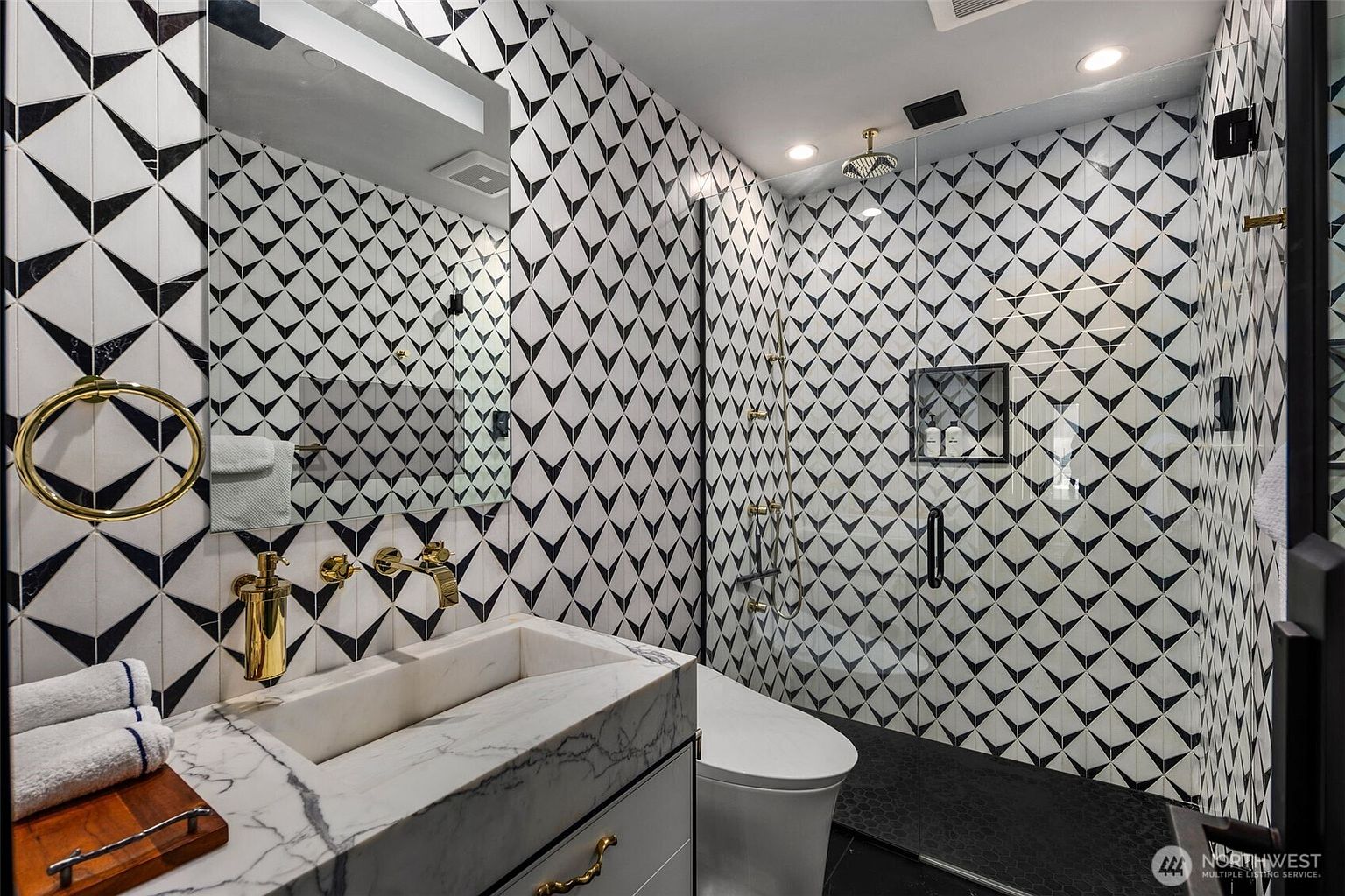 This modern bathroom features a striking geometric patterned tile wall that serves as a bold focal point. The space includes a stone-topped vanity with a rectangular sink, gold-toned fixtures, and a glass-enclosed shower area with a dark pebble-style floor. The perspective captures the vanity and shower in a single frame, highlighting the high-contrast design and luxurious finishes.
