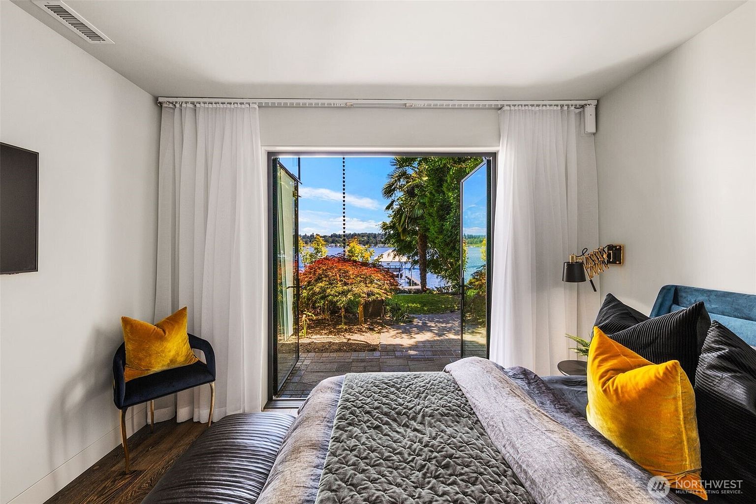 This primary bedroom features a sophisticated design with a plush bed adorned with dark and yellow accent pillows, centered in front of large glass doors that open to a scenic waterfront view. The room is framed by elegant white curtains and dark wood flooring, creating a serene and luxurious atmosphere. The perspective looks directly through the open doors toward a lush garden and a calm lake, emphasizing the seamless indoor-outdoor connection.