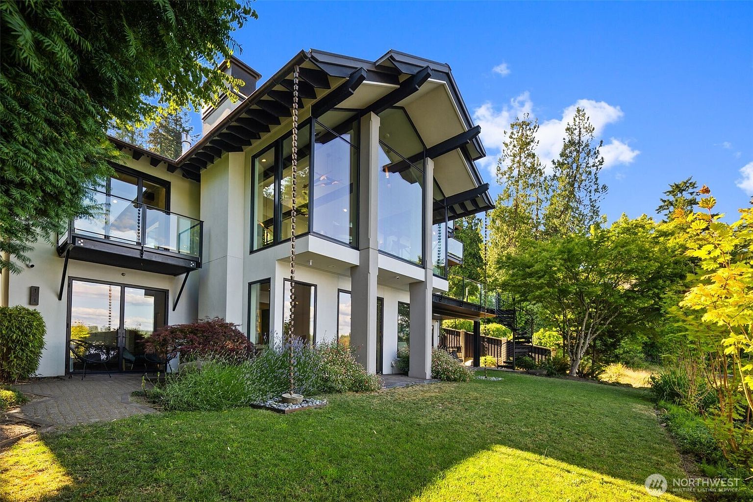 This contemporary residence features a striking architectural design with expansive floor-to-ceiling glass walls that flood the interior with natural light. The exterior is finished in a clean, light-toned stucco, complemented by dark-framed windows and a prominent gabled roofline. A lush green lawn, mature trees, and a stone patio area create a serene backyard setting, while a metal chain rain gutter adds a unique decorative touch.