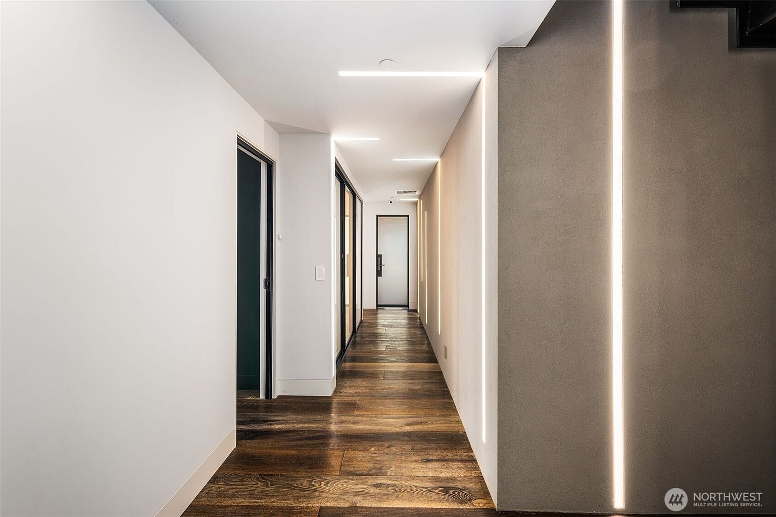 This modern hallway features rich, dark-toned wood flooring and crisp white walls, creating a high-contrast aesthetic. The space is illuminated by sleek, linear recessed lighting integrated into both the ceiling and the side wall, providing a dramatic, architectural glow. The perspective looks down the length of the corridor, highlighting the clean lines and minimalist design that lead toward a glass-paneled door at the far end.