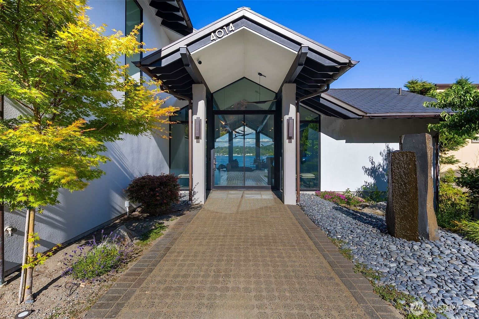 This striking entryway features a modern, angular roofline over a glass-paneled front door, creating a dramatic focal point. A paved walkway leads directly to the entrance, flanked by a vibrant Japanese maple tree on the left and a decorative stone water feature on the right. The perspective is centered and eye-level, emphasizing the clean lines of the architecture and the inviting transition from the exterior to the water view visible through the glass.