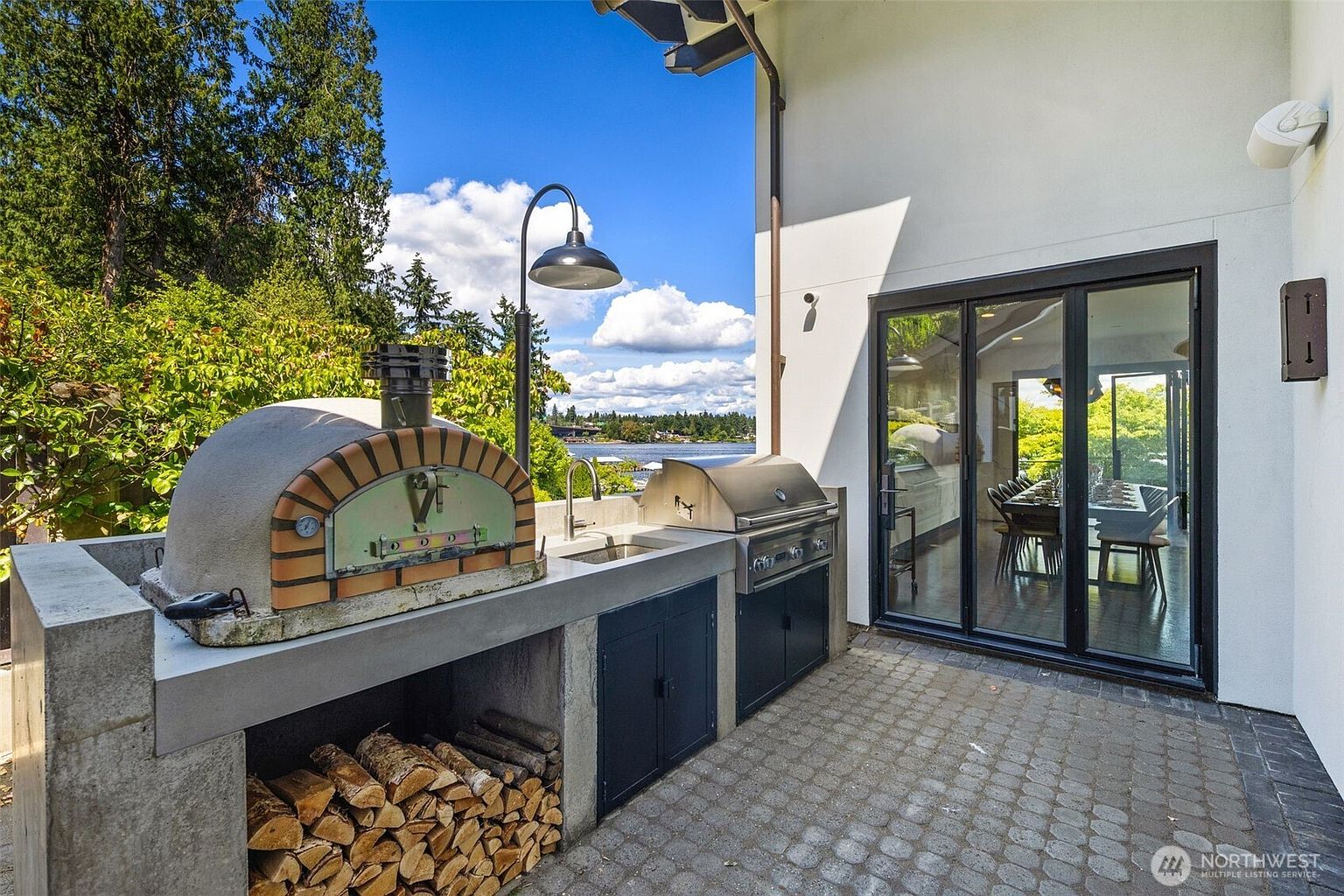 This outdoor patio area features a built-in kitchen setup complete with a stainless steel grill, a sink, and a rustic wood-fired pizza oven. Below the counter, a neatly stacked wood storage area adds to the functional, artisanal aesthetic. The space is paved with stone pavers and offers a scenic view of the water, framed by large glass sliding doors that lead into the home's interior.