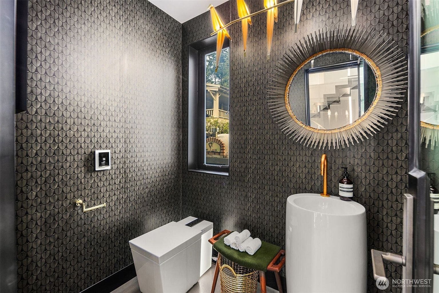 This sophisticated powder room features dramatic, dark patterned wallpaper and a sleek, white pedestal sink with a gold faucet. A striking sunburst-style mirror hangs above the sink, complemented by a unique, sculptural light fixture overhead. The space is finished with a modern toilet and a small wooden bench with rolled towels, creating a high-end, boutique aesthetic.