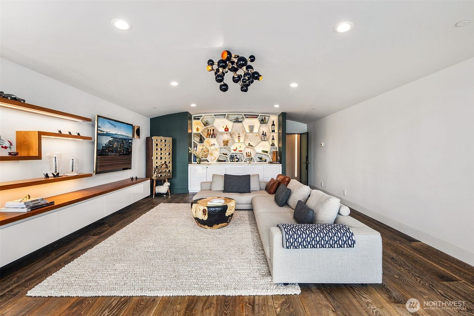 This contemporary living room features a large sectional sofa arranged around a unique, natural-edge wood coffee table on a textured area rug. The space is anchored by a media wall with floating wood shelves and a built-in bar area with geometric shelving and integrated lighting. The room is illuminated by a modern, multi-sphere chandelier and recessed lighting, creating a sophisticated and inviting atmosphere.