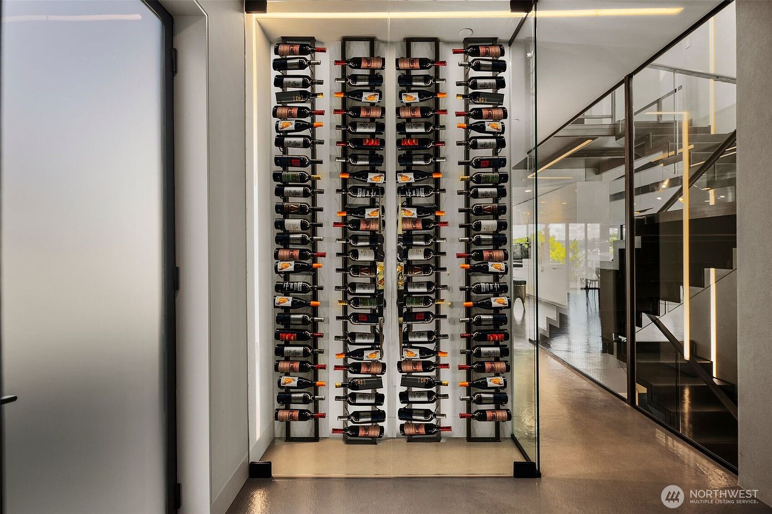 This modern wine cellar features a sleek, glass-enclosed display area with four vertical metal racks holding a curated collection of wine bottles. The minimalist design is complemented by clean lines, neutral walls, and a polished concrete floor, creating a sophisticated and contemporary aesthetic. A glass partition separates the wine display from an adjacent hallway, offering a clear view of the staircase beyond.