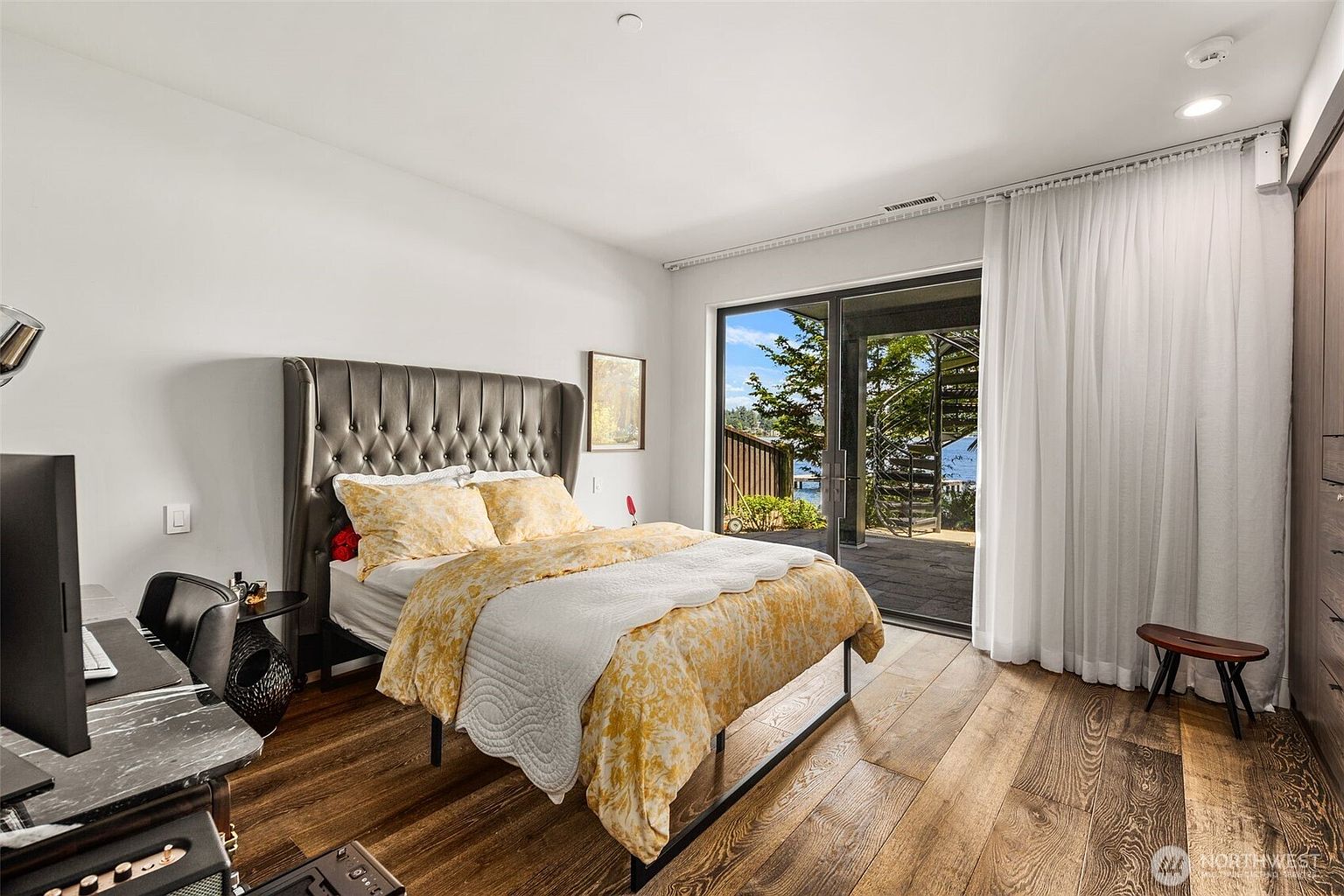 This bright bedroom features a tufted gray headboard and a bed dressed in yellow patterned bedding, set against crisp white walls and warm-toned wood flooring. A large sliding glass door provides access to an outdoor patio with a spiral staircase, allowing for ample natural light and a view of the surrounding greenery. The room is furnished with a sleek desk area to the left and a small wooden stool near the window, creating a functional and inviting living space.