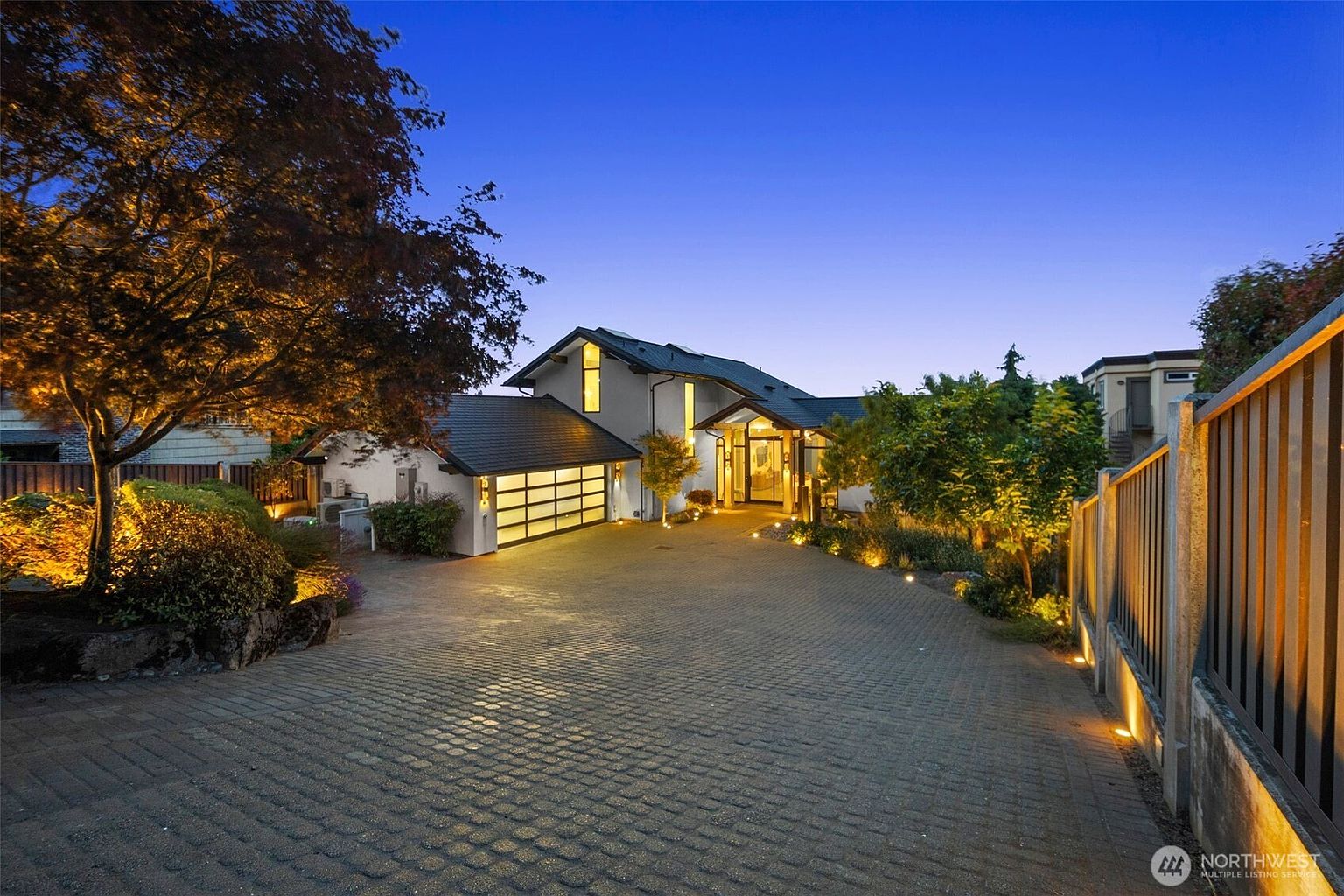 This evening shot captures the front facade of a contemporary home with a paved driveway leading to a multi-car garage featuring frosted glass panels. The house showcases a modern architectural style with clean lines, warm exterior lighting, and a prominent entryway. A large, mature tree frames the left side of the composition, while the twilight sky provides a deep blue backdrop to the illuminated property.