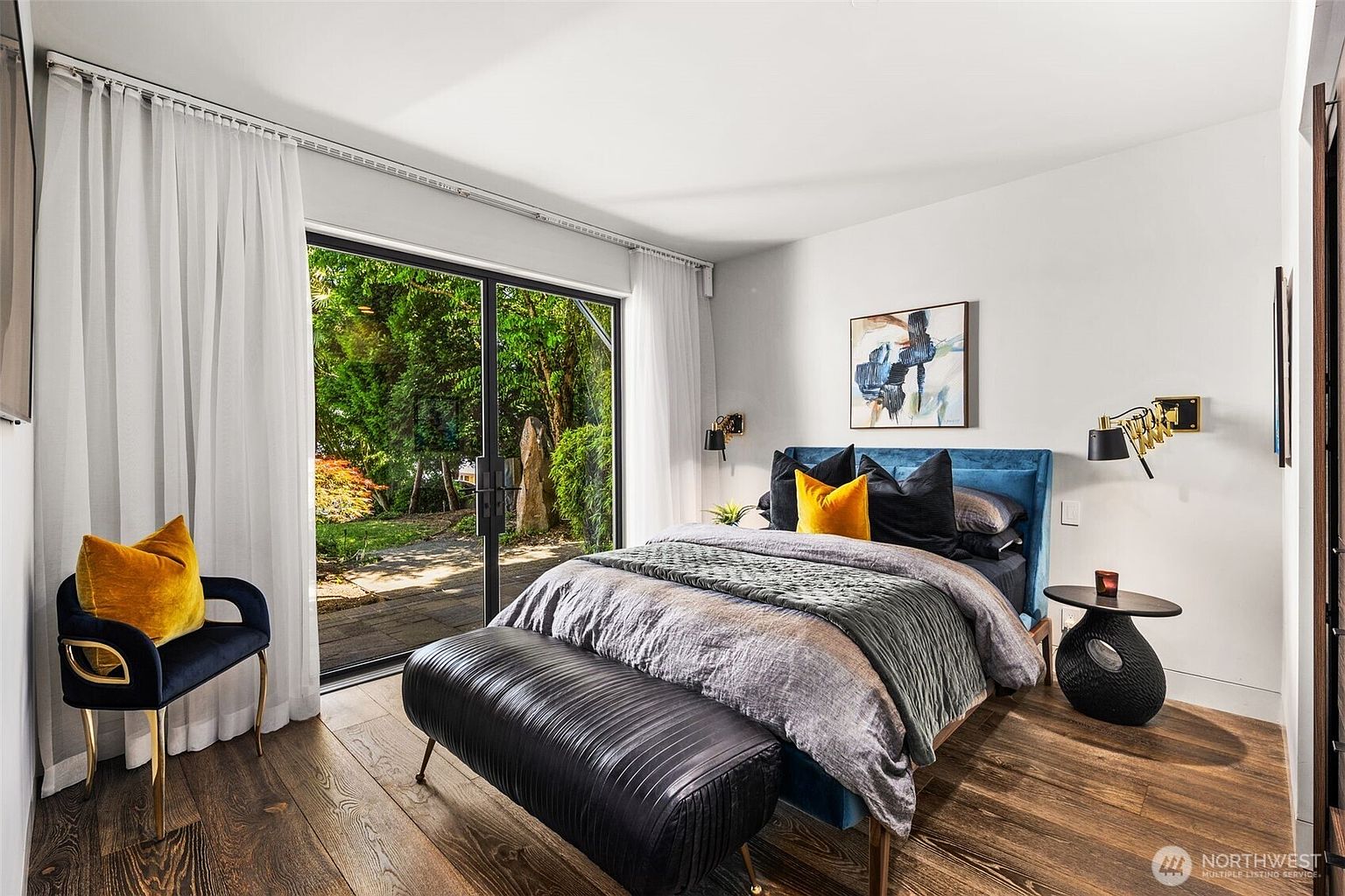 This modern primary bedroom features a plush blue velvet bed frame accented with gold and yellow throw pillows, positioned against a crisp white wall. A sleek black bench sits at the foot of the bed, while large sliding glass doors provide a seamless transition to the lush, green outdoor garden space. The room is finished with warm wood flooring and contemporary wall-mounted lighting, creating a sophisticated and tranquil atmosphere.