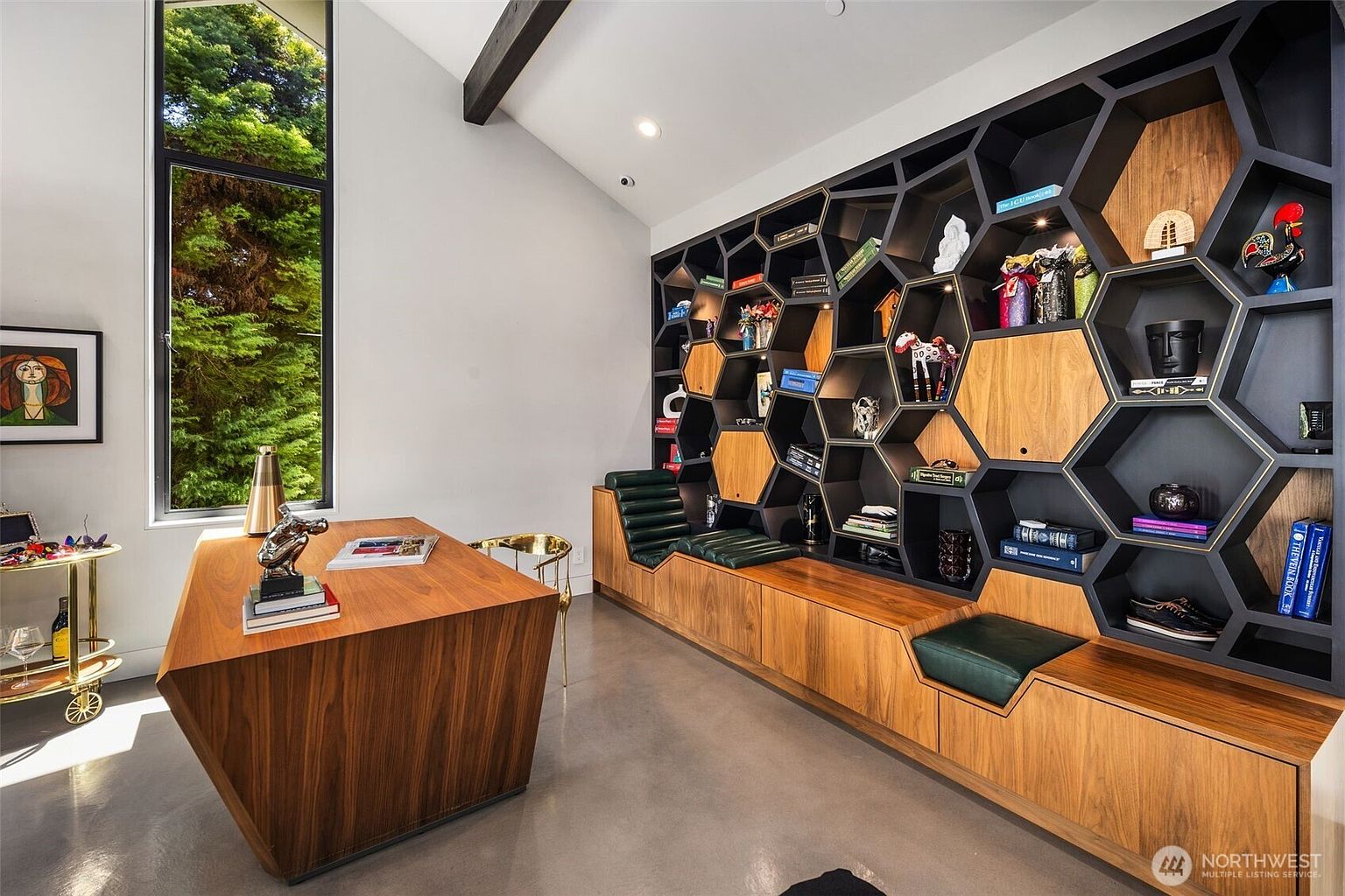 This modern home office features a striking, geometric honeycomb-style shelving unit that spans the wall and incorporates built-in bench seating with dark green cushions. A large, angular wood desk sits in the center of the room, complemented by a tall window that provides ample natural light and a view of the surrounding greenery. The space is finished with polished concrete floors and a minimalist aesthetic, creating a sophisticated and productive atmosphere.