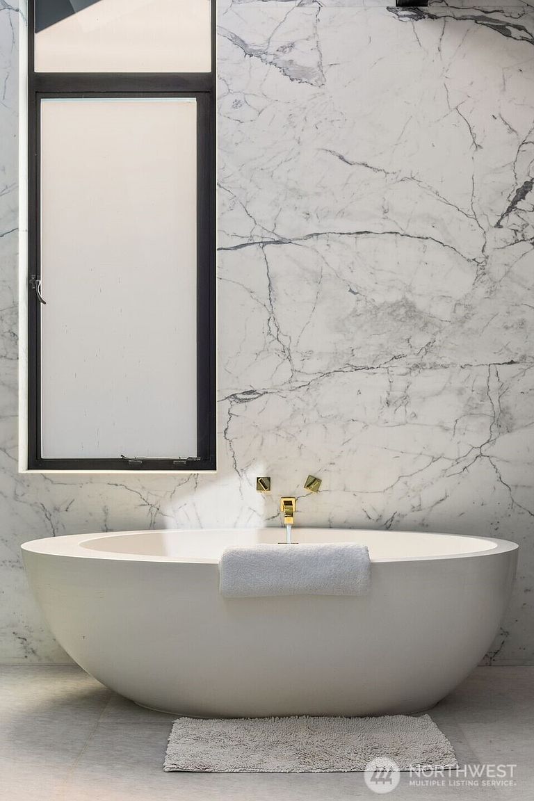 This luxurious primary bathroom features a deep, oval-shaped soaking tub centered against a dramatic wall clad in large-format, veined stone slabs. A tall, rectangular window with a dark frame provides natural light, while gold-toned fixtures add a touch of elegance to the minimalist design. A soft bath mat and a folded towel on the tub edge complete the serene, spa-like atmosphere.
