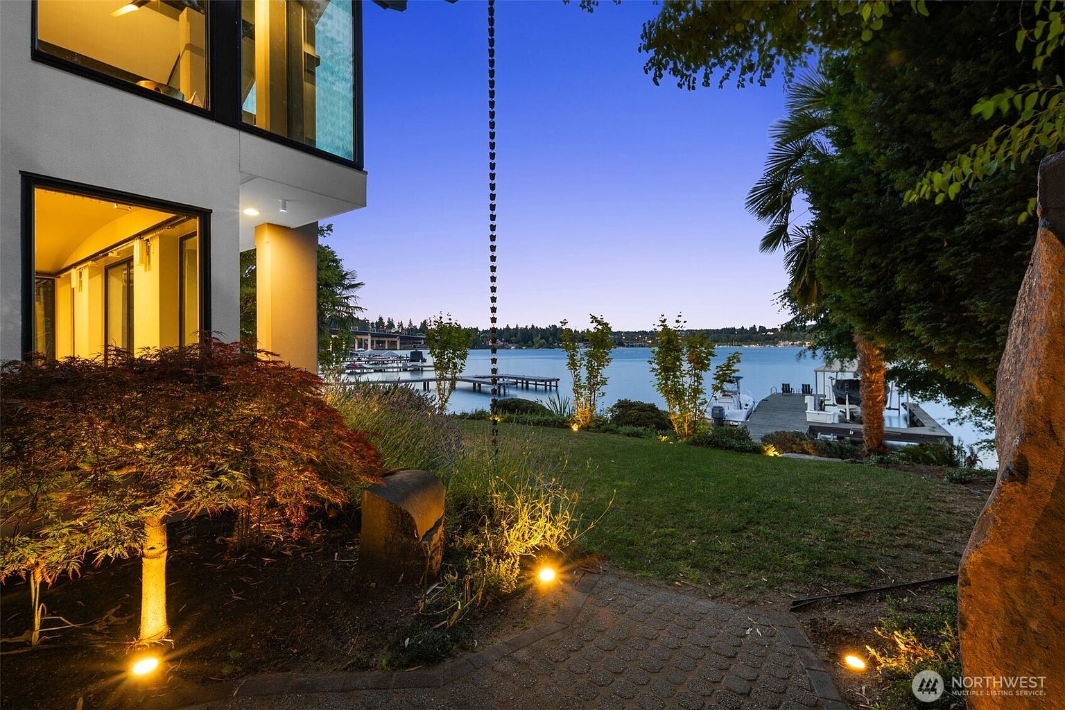 This evening view captures the rear of a modern residence overlooking a calm lake, featuring large windows that glow with warm interior light. A manicured lawn leads down to a private dock and boat slip, framed by mature trees and soft landscape lighting. The perspective is from a paved garden path, emphasizing the seamless transition between the home's architecture and the waterfront setting.