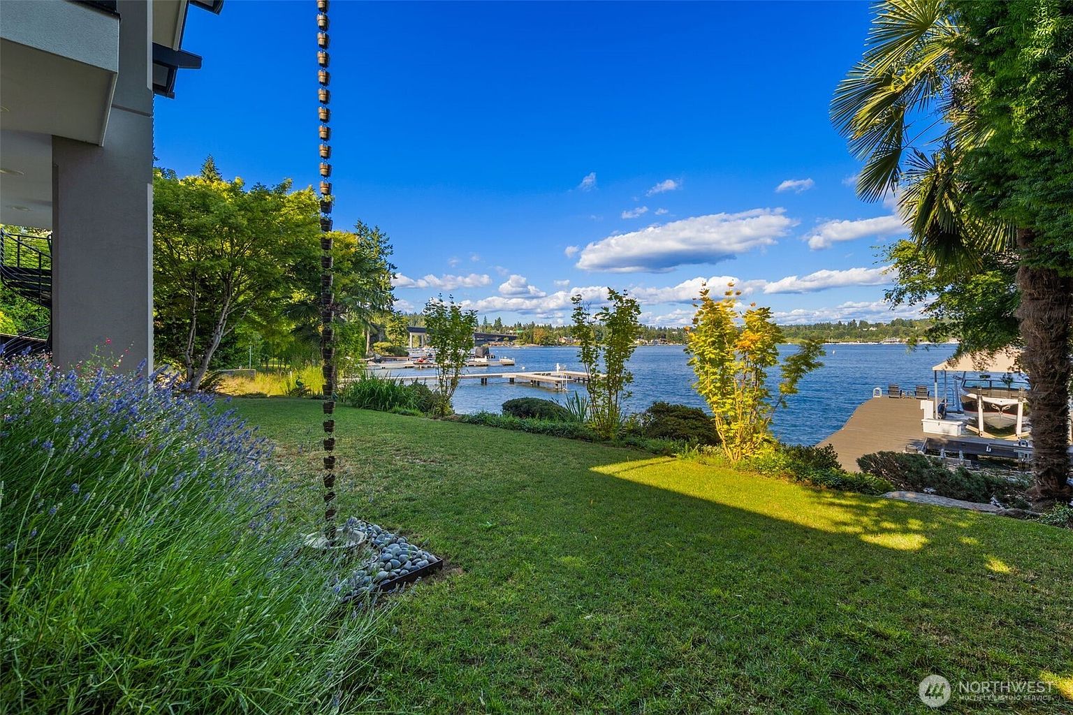 This scenic backyard view captures a lush green lawn leading directly to a private waterfront with a dock and boat lift. The perspective from the side of the house highlights a decorative rain chain hanging near a garden bed filled with lavender, while mature trees and a palm tree frame the expansive lake vista under a bright, cloudy sky. The setting offers a tranquil and luxurious outdoor living space perfect for waterfront recreation.