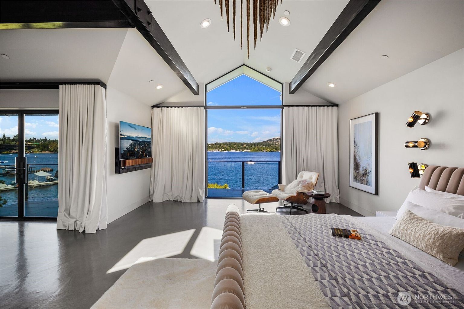 This luxurious primary bedroom features a vaulted ceiling with exposed dark beams and a striking chandelier, creating a dramatic architectural focal point. A large glass wall provides an expansive, unobstructed view of the lake and private balcony, while a modern lounge chair and ottoman offer a serene seating area. The room is finished with polished concrete floors, a plush bed with a patterned quilt, and contemporary wall-mounted lighting fixtures.