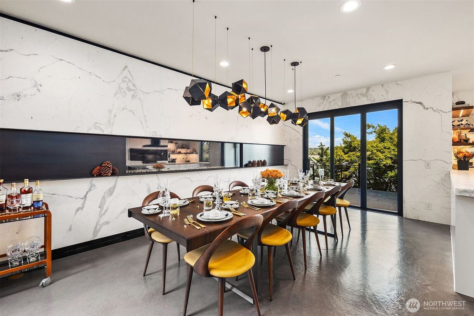 This sophisticated dining room features a long, dark wood table surrounded by mid-century modern chairs with yellow upholstered seats. A striking, geometric multi-pendant light fixture hangs above the table, while a long, horizontal recessed shelf is set into a wall clad in white stone with grey veining. Large sliding glass doors provide a clear view of the outdoors, and a bar cart sits to the left, completing the elegant and contemporary aesthetic.