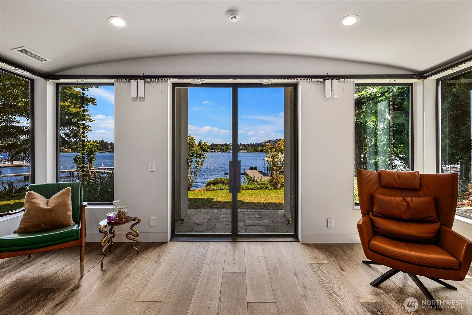 This bright sunroom features light wood flooring and large, expansive windows that frame a serene lake view. The space is furnished with a green armchair on the left and a tan leather swivel chair on the right, centered around a sliding glass door that leads to a patio. The room has a clean, modern aesthetic with recessed lighting and a neutral color palette that emphasizes the natural scenery outside.