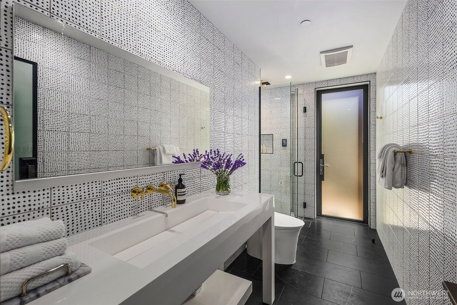 This modern bathroom features a striking, high-contrast aesthetic with patterned, grid-like wall tiles and dark, large-format floor tiles. A long, white vanity with a double-faucet sink and gold-toned hardware serves as the focal point, complemented by a large, frameless mirror and a vase of fresh lavender. The space includes a glass-enclosed shower area and a sleek, minimalist toilet, creating a sophisticated and contemporary atmosphere.