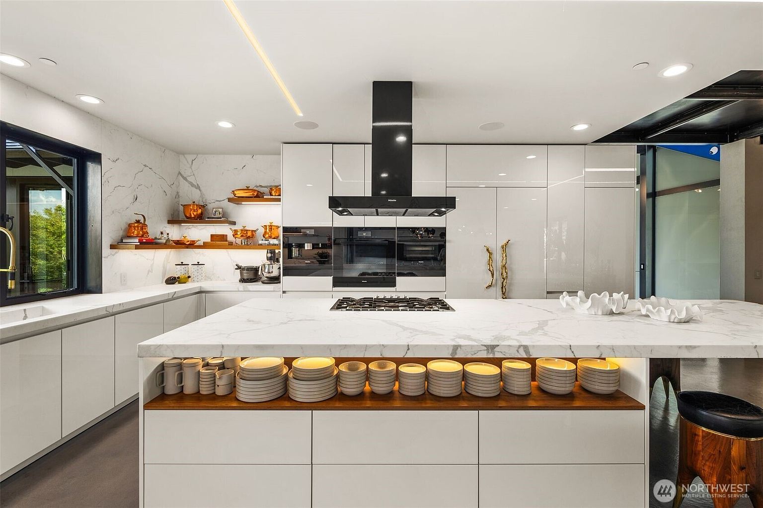 This modern, high-end kitchen features a large central island with a stone countertop and integrated open shelving displaying stacks of white plates. The space is equipped with sleek, glossy white cabinetry, a prominent black range hood, and built-in appliances. Warm under-cabinet lighting and recessed ceiling lights create an inviting atmosphere, while open wooden shelves on the left wall provide a decorative touch.