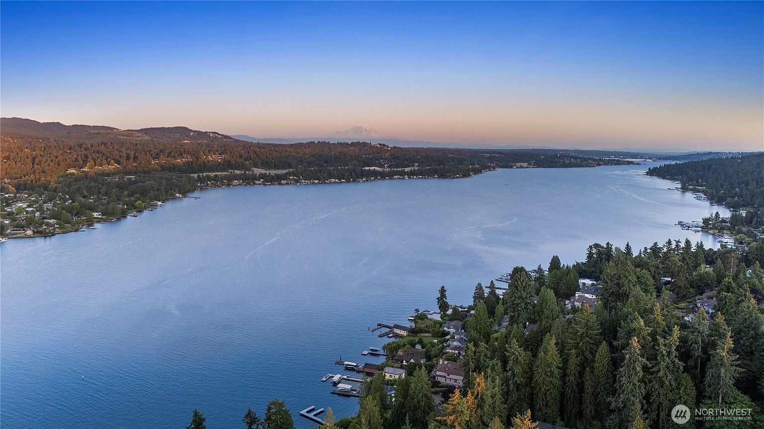 This high-angle aerial view captures a serene lakefront setting, showcasing a vast expanse of water surrounded by lush, dense evergreen forests and residential properties. In the distance, a prominent snow-capped mountain peak rises above the horizon, providing a dramatic backdrop to the tranquil landscape. The perspective emphasizes the expansive nature of the waterfront community and the peaceful, natural environment.