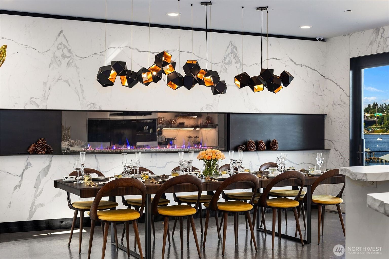 This sophisticated dining room features a long, dark-toned table surrounded by mid-century modern style chairs with mustard-yellow upholstery. A striking, geometric multi-pendant light fixture hangs above the table, set against a dramatic wall with a long, linear fireplace integrated into a large-format stone panel. The space is elegantly staged with place settings and a floral centerpiece, creating a luxurious and inviting atmosphere for formal dining.