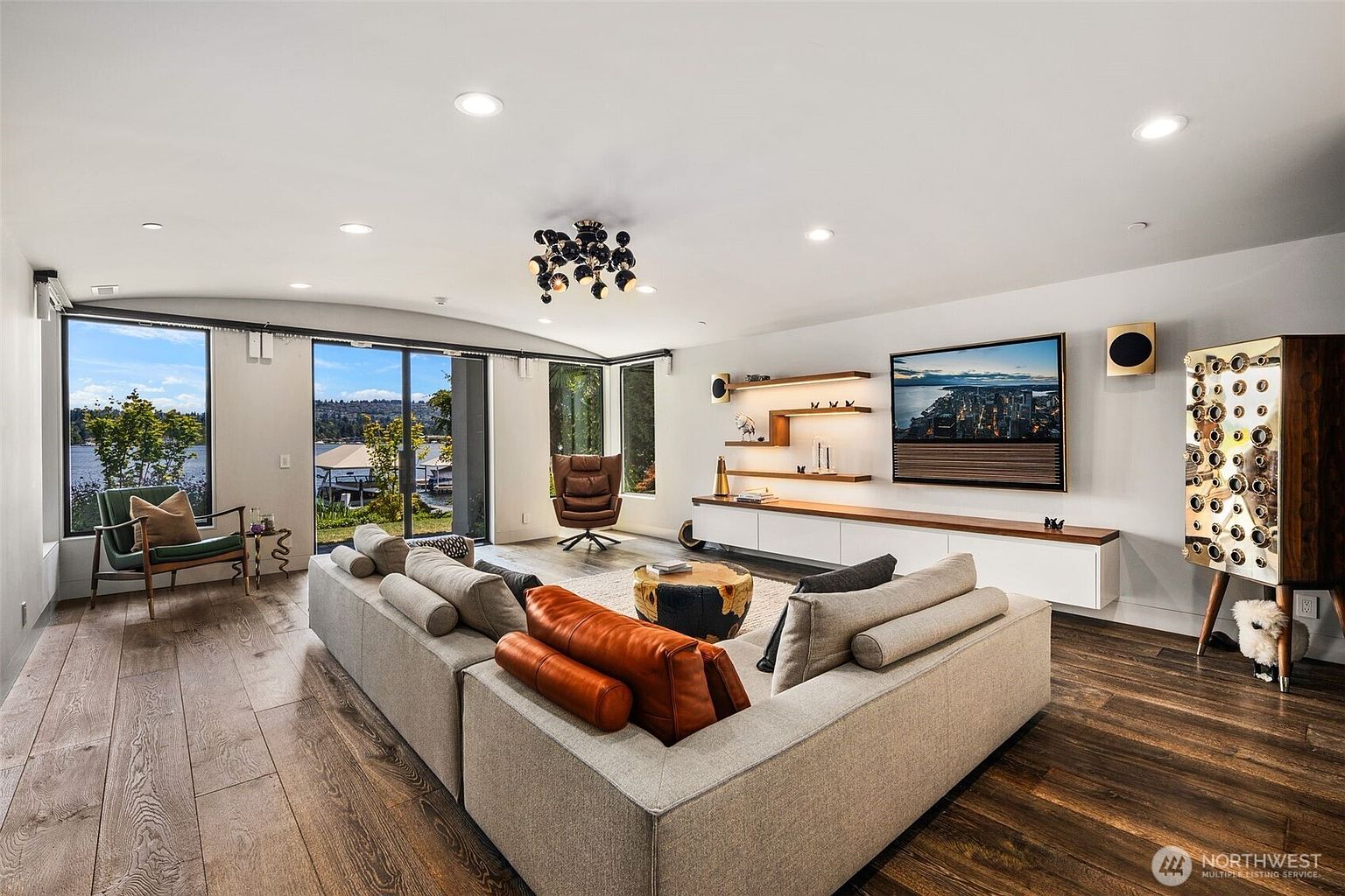 This spacious, modern living room features a large sectional sofa with leather accents, a central coffee table, and a sleek media console mounted below a wall-mounted television. The room is illuminated by a unique, multi-bulb chandelier and recessed lighting, while large sliding glass doors provide an expansive view of the lake outside. The interior design combines warm wood flooring with a neutral color palette, creating a sophisticated and inviting atmosphere.