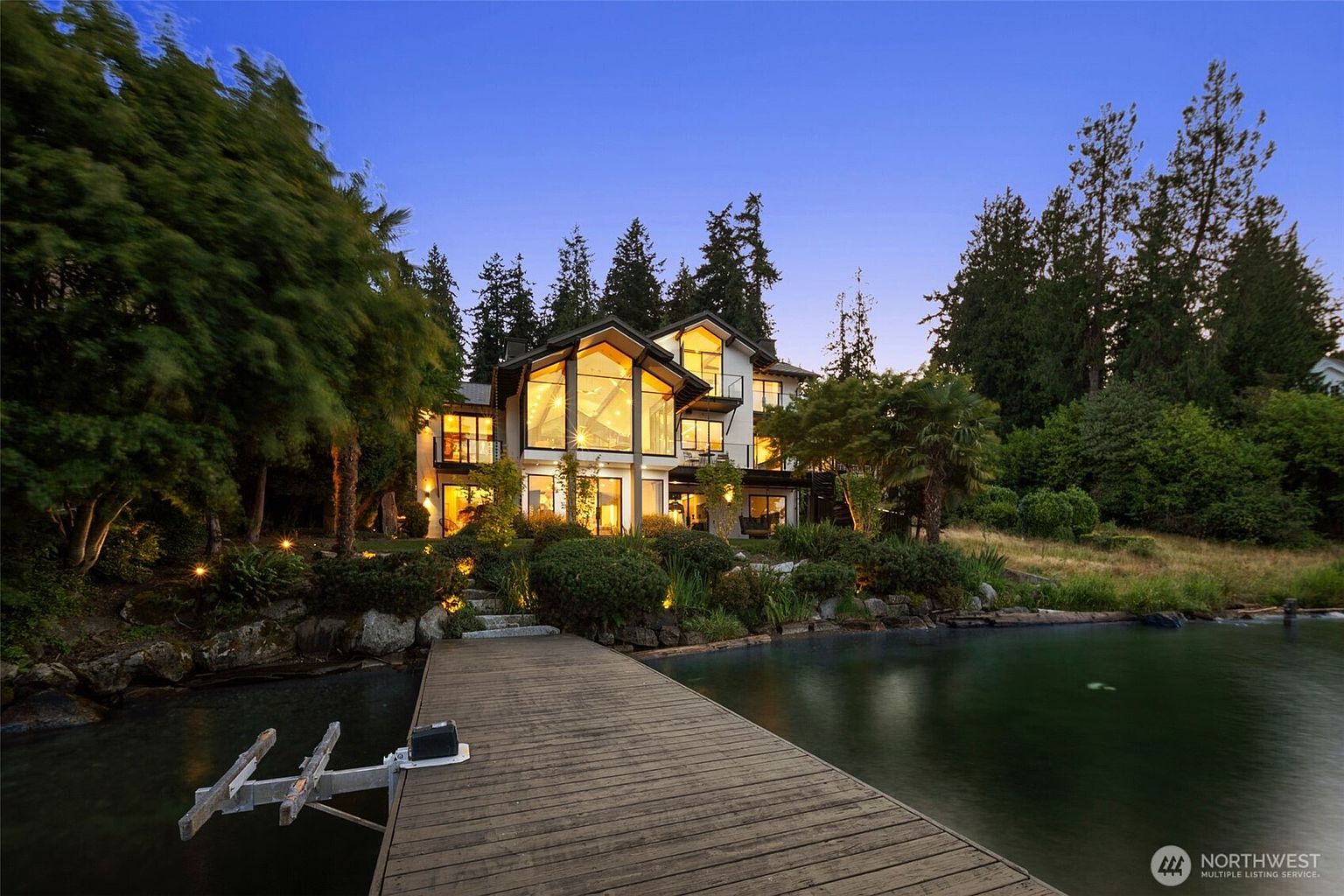 This stunning waterfront residence is captured at dusk, showcasing a multi-level architectural design with expansive glass walls that illuminate the interior. A wooden dock extends from the foreground into the calm water, leading the eye toward the landscaped yard and the home's prominent, angular roofline. The setting is serene and private, surrounded by mature trees and lush greenery, creating a sophisticated and inviting atmosphere.