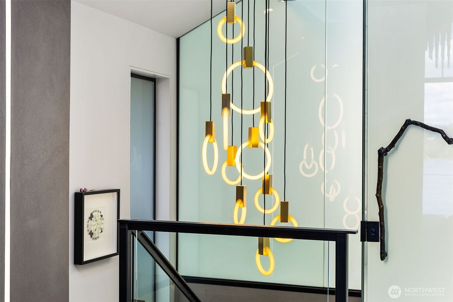 This interior view captures a sophisticated staircase landing featuring a striking, multi-tiered gold ring chandelier that serves as a focal point. To the right, a frosted glass partition provides privacy while allowing light to filter through, complemented by a decorative branch-like sculpture mounted on the wall. The space is defined by a clean, modern aesthetic with a black metal railing and a framed piece of art on the adjacent wall, creating an elegant and artistic transition area.