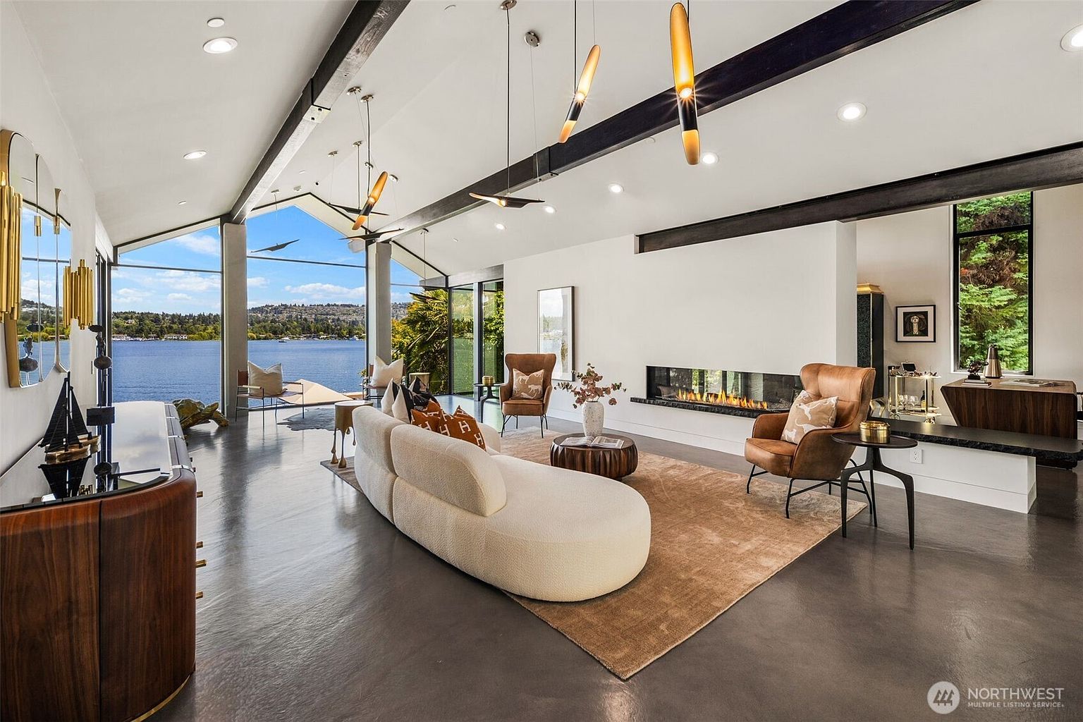 This expansive, modern living room features a curved sofa and leather armchairs arranged on a large area rug over polished concrete floors. A long, linear fireplace is set into a white wall, while floor-to-ceiling windows provide a panoramic view of the lake and surrounding landscape. The room is accented by dark exposed ceiling beams and unique pendant lighting, creating a sophisticated and airy atmosphere.