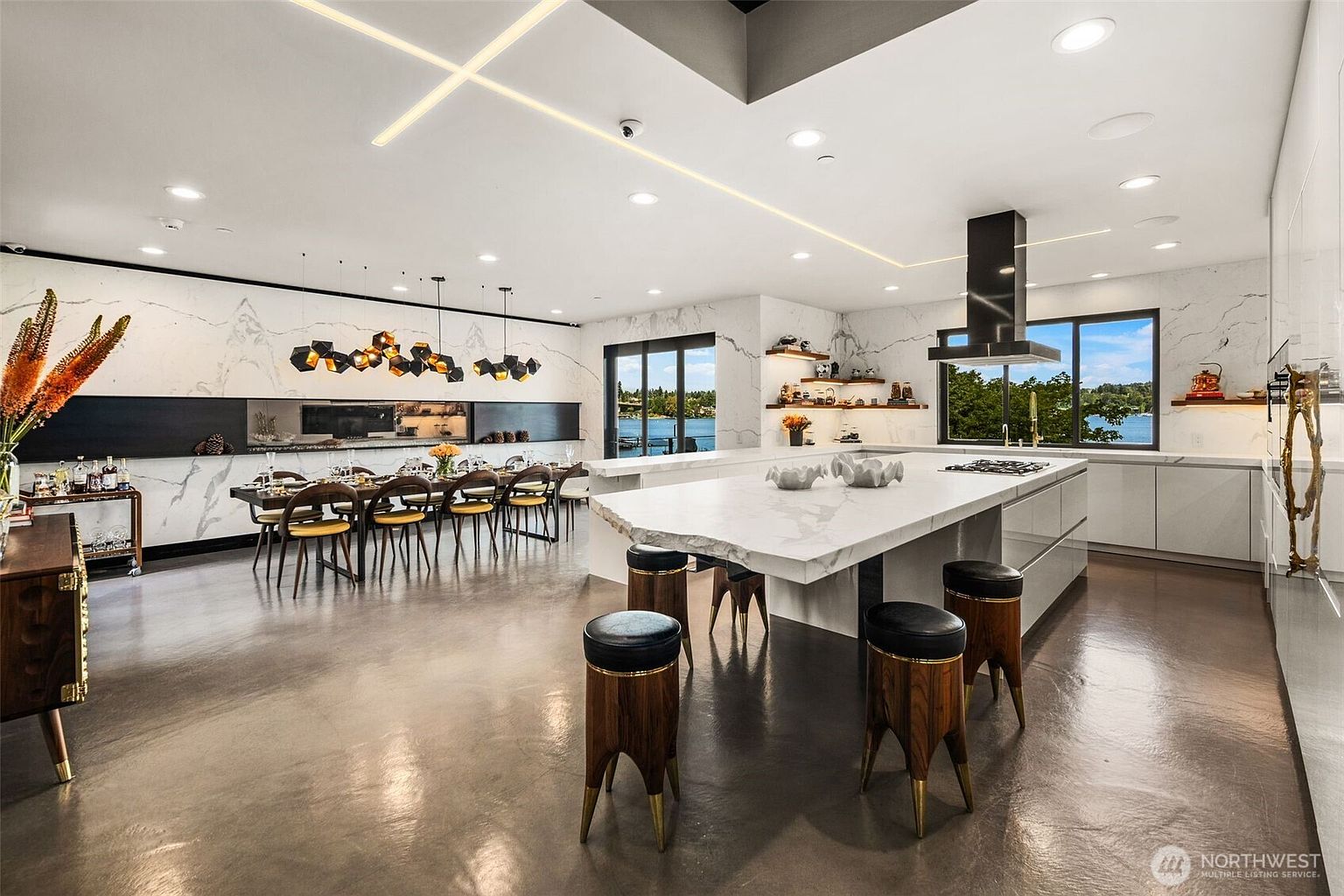 This expansive, open-concept kitchen and dining area features a large central island with stone countertops and unique sculptural stools. The space is defined by sleek, minimalist cabinetry, a professional-grade range hood, and a long dining table illuminated by geometric pendant lights. Large windows provide a scenic view of the water, while the polished concrete floors and white stone wall accents create a sophisticated, modern aesthetic.