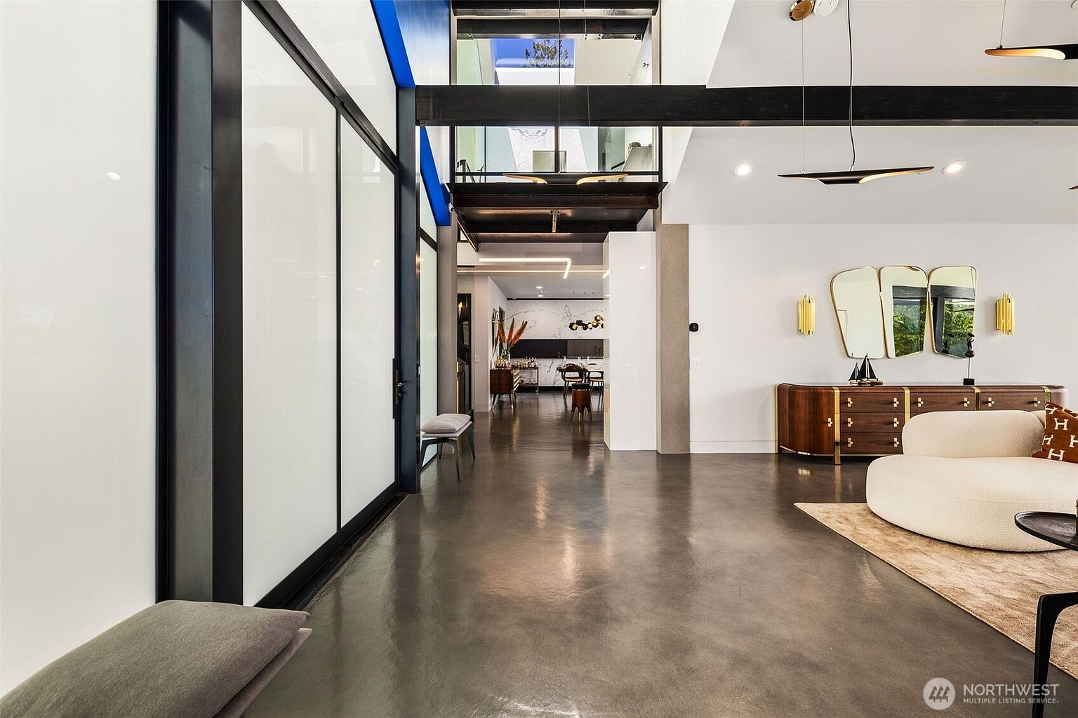 This expansive, modern living space features polished concrete floors and a dramatic open-concept layout with a high, vaulted ceiling. A large, curved white sofa sits on a textured rug, while a long wooden sideboard with gold hardware is accented by a trio of decorative mirrors. The perspective looks through the main living area toward a bright, open hallway that leads to a dining space, showcasing a sophisticated, architectural design with clean lines and high-end finishes.