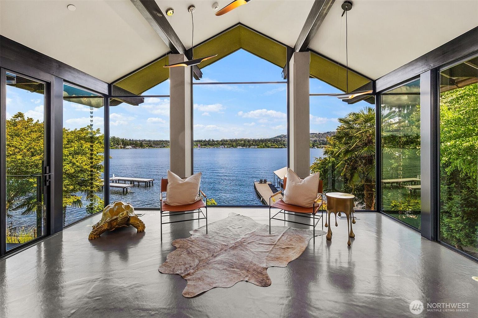 This sun-drenched room features expansive floor-to-ceiling glass walls that frame a panoramic view of the lake and private dock. The space is anchored by a polished concrete floor topped with a cowhide rug, two leather chairs, and a decorative gold-toned turtle sculpture. The vaulted ceiling with exposed beams and modern pendant lighting creates an airy, sophisticated atmosphere perfect for relaxation.