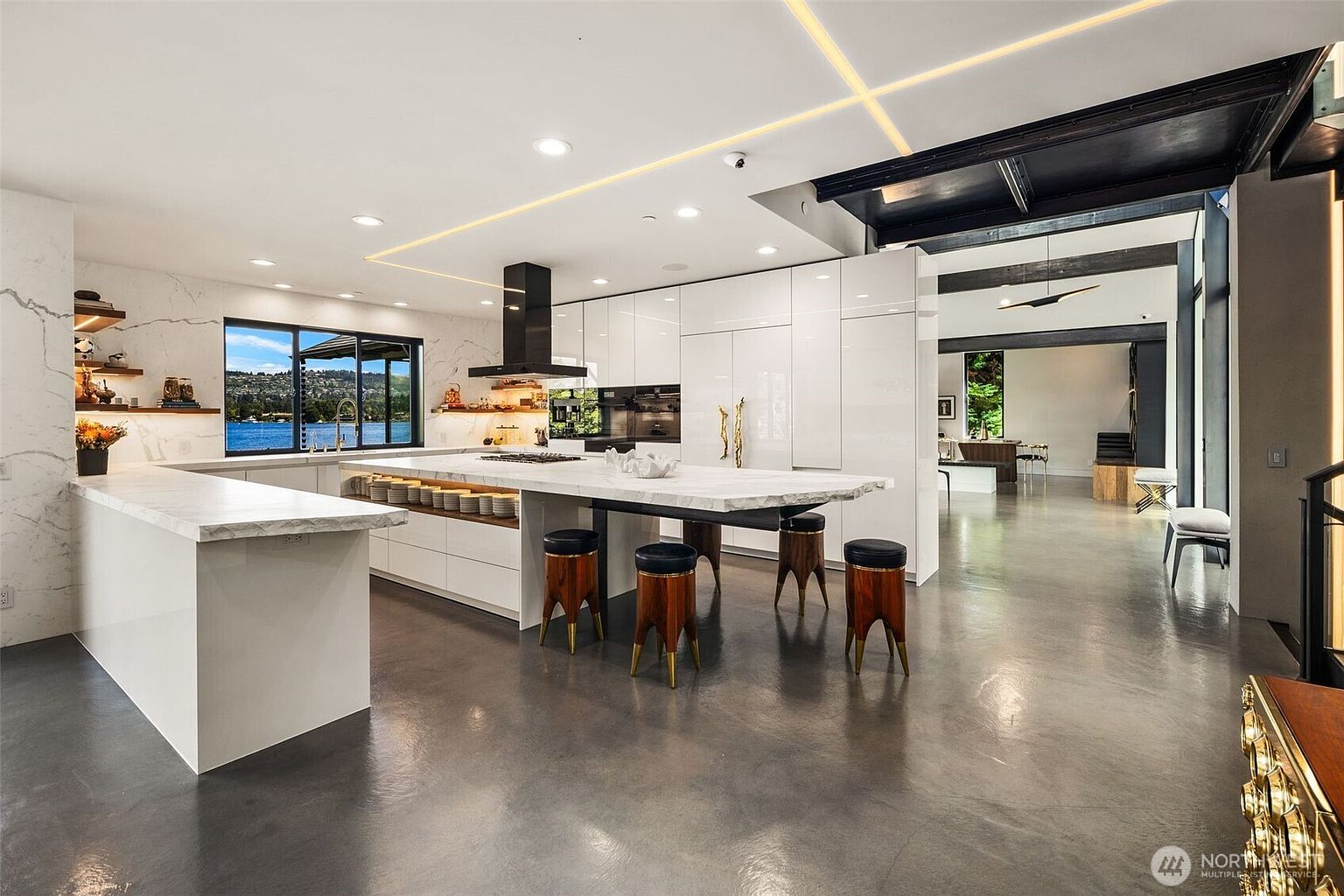 This expansive, modern kitchen features a large central island with a stone countertop and integrated seating, complemented by sleek white cabinetry and high-end appliances. The space is defined by polished concrete floors, minimalist lines, and a wide window that frames a scenic view of the water. The open-concept design flows seamlessly into an adjacent living area, creating a bright and sophisticated atmosphere.