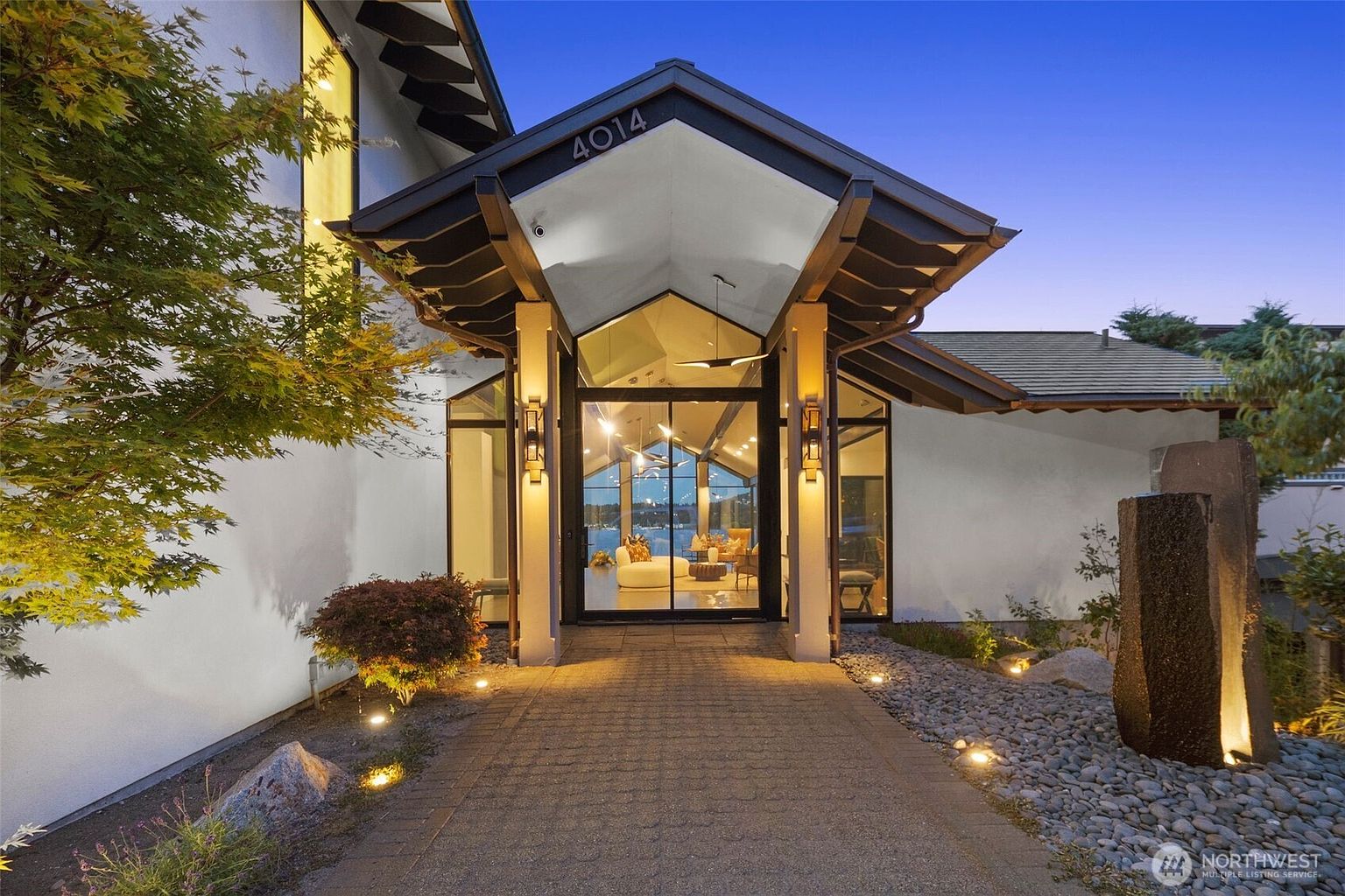This striking modern entryway features a dramatic, angular roofline over a paved walkway leading to a glass-fronted entrance. The home is illuminated by warm, recessed lighting that highlights the architectural geometry and the surrounding landscaping, including a stone water feature. The perspective is a low-angle, eye-level shot that emphasizes the grand scale and inviting nature of the home's facade at dusk.
