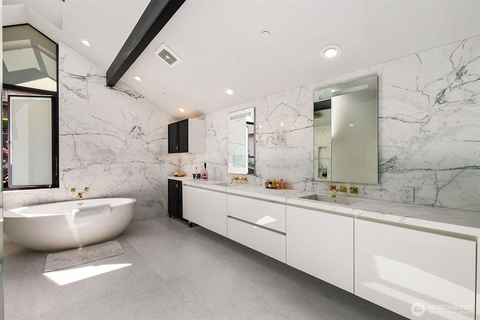 This luxurious primary bathroom features extensive stone wall cladding with dramatic veining, creating a sophisticated and high-end aesthetic. The space includes a sleek, white double vanity with gold-toned fixtures, a freestanding soaking tub, and a minimalist design palette. Large windows and recessed lighting brighten the room, which is finished with neutral-toned flooring.