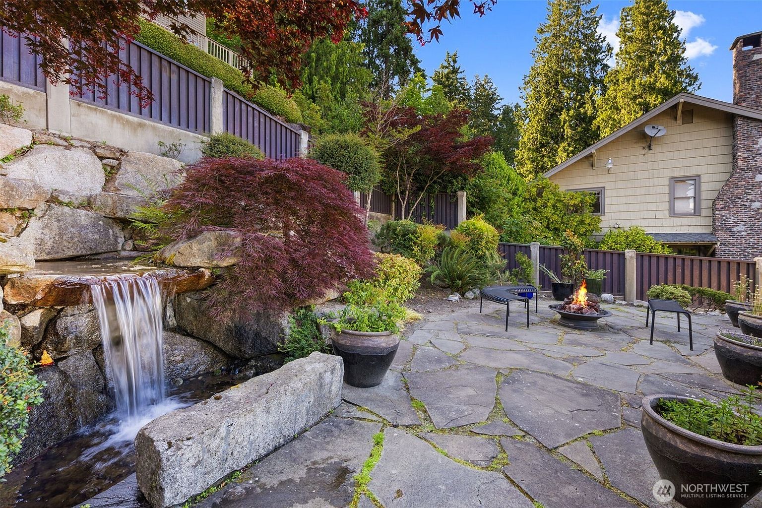 This inviting outdoor living space features a flagstone patio centered around a fire pit, complemented by a custom rock waterfall feature. The area is surrounded by lush landscaping, including a prominent Japanese maple, and is enclosed by a privacy fence. The perspective captures a serene backyard retreat, highlighting the blend of natural stone elements and manicured greenery.