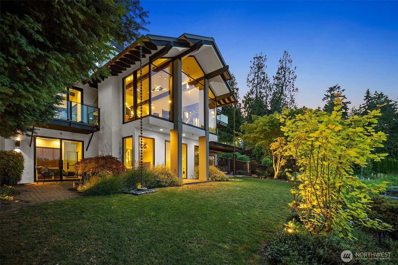 This striking contemporary home features a dramatic two-story glass facade that floods the interior with light and offers a seamless connection to the outdoors. The exterior is characterized by clean lines, dark structural framing, and multiple balconies, set against a lush, manicured lawn at twilight. Warm exterior lighting highlights the landscaping and the architectural geometry of the building, creating an inviting and sophisticated atmosphere.