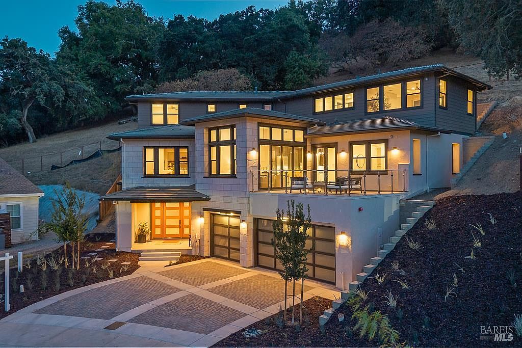 This striking contemporary home features a multi-level design with a blend of stone and dark siding, set against a hillside backdrop. The front facade showcases a prominent entryway with a warm wood door, a spacious elevated deck, and a two-car garage with modern glass-paneled doors. The paved driveway and manicured landscaping create an inviting and sophisticated curb appeal, captured during the golden hour.