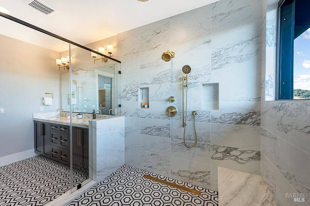 This luxurious primary bathroom features a spacious walk-in shower with floor-to-ceiling marble tile, gold-toned fixtures, and a built-in bench. The vanity area is separated by a glass partition, showcasing dark cabinetry and a patterned tile floor that adds a modern, sophisticated touch. The overall aesthetic is high-end and contemporary, emphasizing clean lines and premium materials.