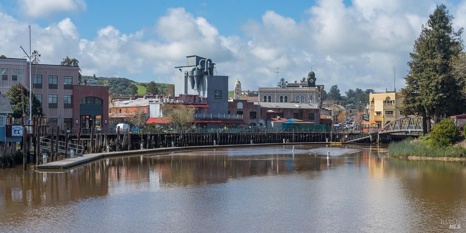 This scenic, eye-level shot captures the Petaluma River waterfront, showcasing a blend of historic industrial architecture and charming downtown buildings. The composition features a calm, reflective river in the foreground, leading the eye toward a prominent mill structure and a pedestrian bridge on the right. The bright, partly cloudy sky and lush greenery create an inviting, picturesque atmosphere characteristic of a vibrant riverfront community.