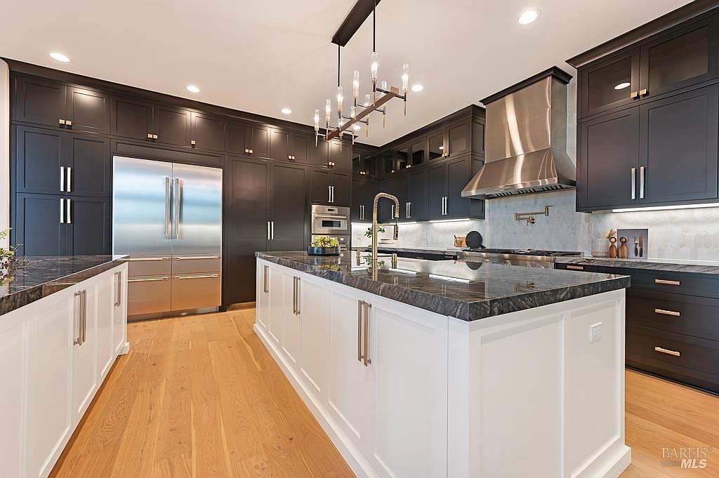 This sophisticated, high-end kitchen features a striking contrast between dark charcoal cabinetry and a crisp white central island, topped with luxurious dark stone countertops. The space is equipped with professional-grade stainless steel appliances, including a large refrigerator and a prominent range hood, all illuminated by a modern linear chandelier. The warm hardwood flooring balances the dramatic color palette, creating an inviting and elegant atmosphere perfect for culinary enthusiasts.