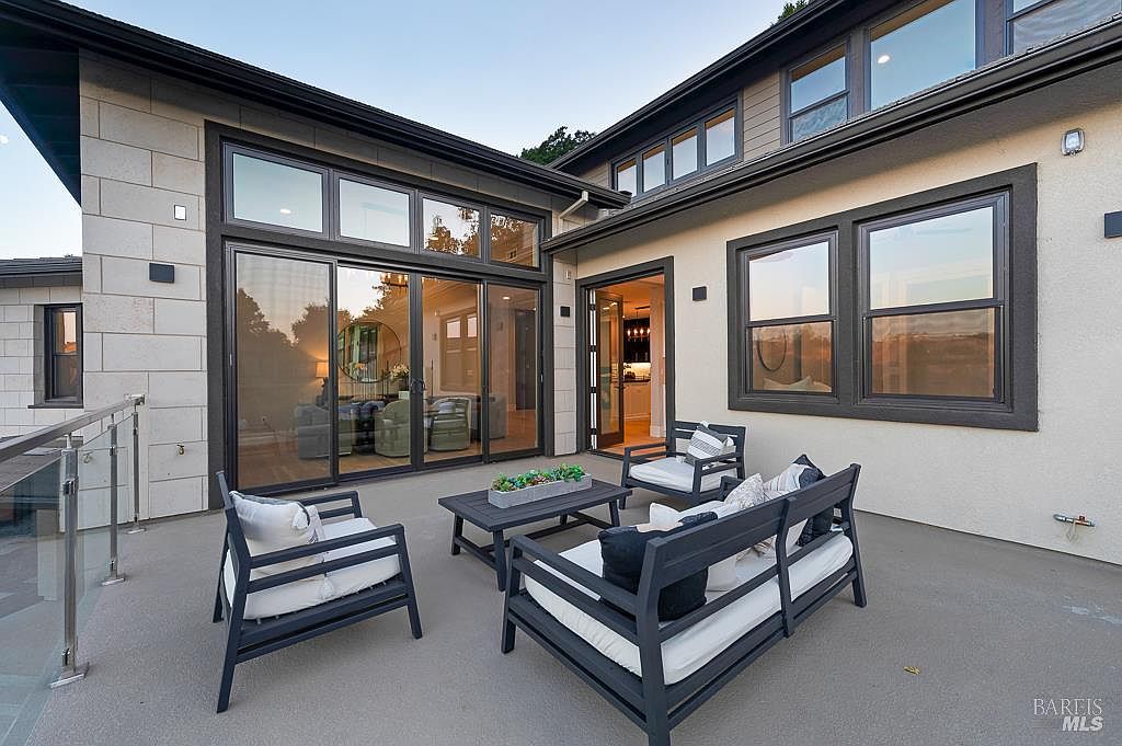 This modern, elevated patio features sleek dark-framed outdoor furniture with plush white cushions, centered around a low-profile coffee table with a succulent centerpiece. The space is framed by large glass sliding doors and windows that offer a seamless transition to the interior, while a glass railing provides an unobstructed view. The overall aesthetic is contemporary and sophisticated, perfect for outdoor entertaining.