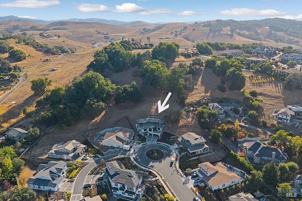 This high-angle aerial view captures a suburban neighborhood nestled against rolling, sun-drenched hills. A prominent white arrow points toward a modern multi-story residence situated at the end of a cul-de-sac, surrounded by other upscale homes and mature trees. The perspective emphasizes the property's integration into the landscape and its quiet, elevated location.