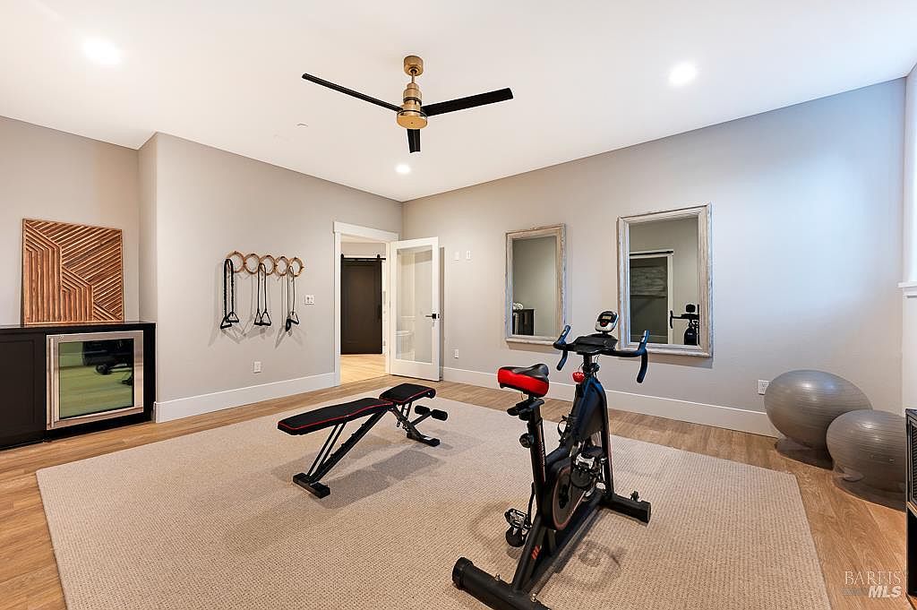 This well-lit home gym features a neutral color palette, light wood flooring, and a large area rug, creating a clean and inviting workout space. The room is equipped with a stationary exercise bike, an adjustable weight bench, exercise balls, and wall-mounted resistance bands, complemented by two large mirrors and a sleek ceiling fan. A modern black cabinet with a glass-front beverage cooler adds a touch of luxury to this functional fitness area.