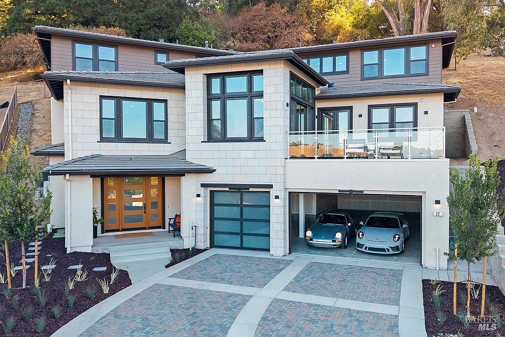 This modern, multi-level luxury residence features a sophisticated blend of light-colored stone masonry and dark wood-toned siding, creating a striking contemporary aesthetic. The front facade includes a prominent entryway with a warm wood-paneled door, a spacious two-car garage housing two classic sports cars, and an elevated balcony with a sleek glass railing. The property is set against a hillside backdrop with a neatly landscaped front yard and a paved driveway, conveying an impression of upscale, refined living.