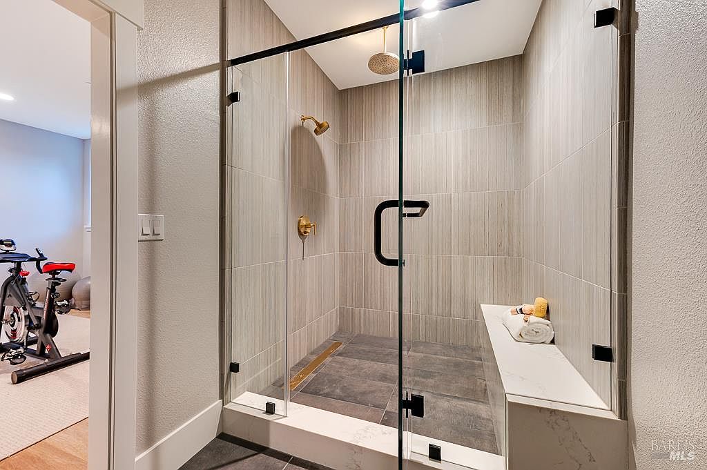 This modern walk-in shower features large-format, light-toned vertical wall tiles and dark, textured floor tiles, creating a sophisticated contrast. It includes a built-in marble bench, a gold-finished rainfall showerhead, and a sleek glass enclosure, with a glimpse of an adjacent home gym visible through the doorway. The overall aesthetic is clean, luxurious, and contemporary, emphasizing high-end finishes and thoughtful design.