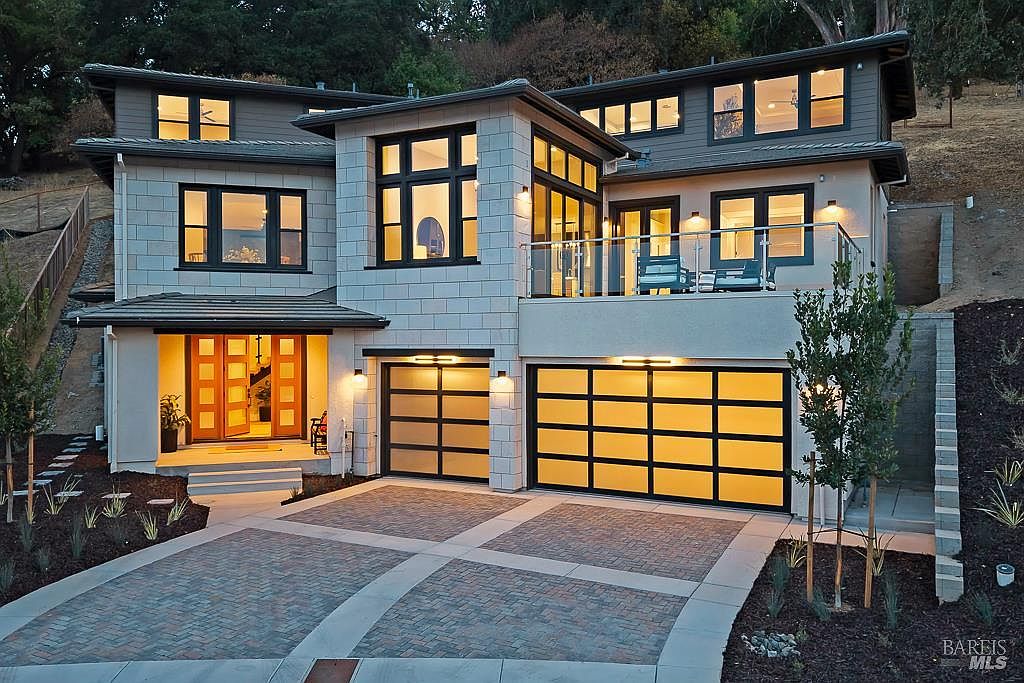 This striking contemporary home features a multi-level facade with a blend of light-colored stone masonry and dark-trimmed windows, creating a sophisticated modern aesthetic. The property includes a spacious two-car garage with frosted glass panels, a welcoming front entryway with warm wood-framed doors, and an elevated balcony overlooking the driveway. The paved driveway and manicured landscaping enhance the home's curb appeal, presenting a luxurious and inviting atmosphere.